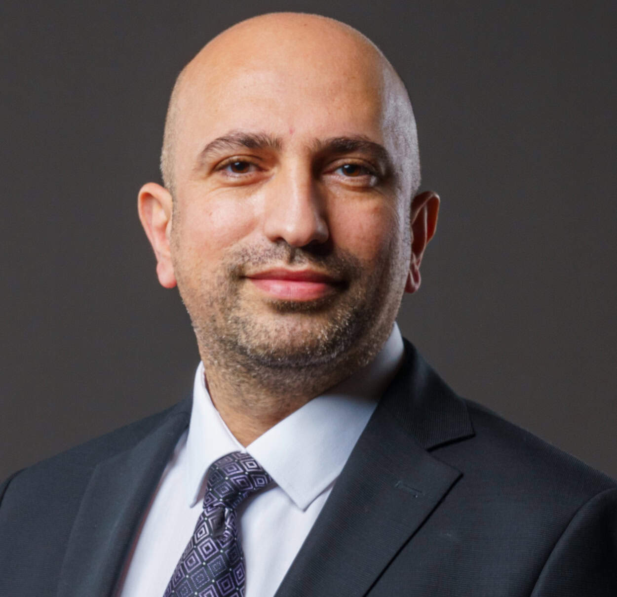 Fatih Kazan - 01 A bald man with light skin and a neatly trimmed beard wears a dark suit, white shirt, and patterned tie, smiling slightly against a plain dark background.